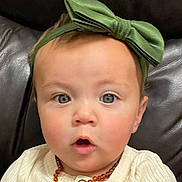 Lillian is registered to the contest to win money with this photo: baby, child, face, headband, green_bow, blue_eyes, amber_necklace, expression, surprised, clothing, buttoned_shirt, infant, portrait, cute, indoor, leather_background, closeup, skin, person, young