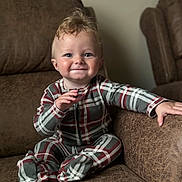Ernie is registered to the contest to win money with this photo: baby, child, plaid_pajamas, smile, happy, couch, brown_furniture, indoor, seated, cute, face, hand, footed_pajamas, young_child, person, cozy, home, infant, expression, portrait