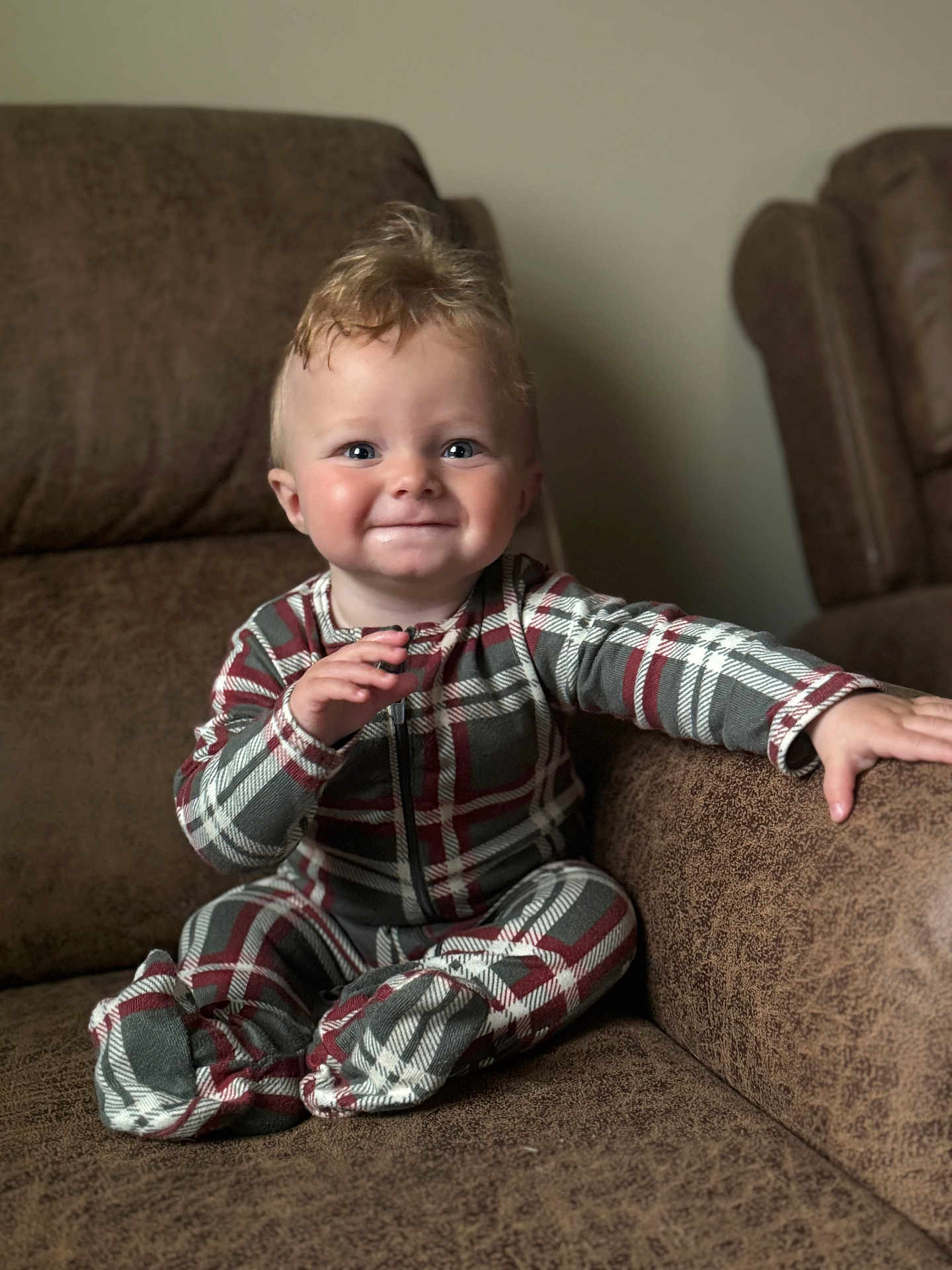 Ernie is registered to the contest to win money with this photo: baby, child, plaid_pajamas, smile, happy, couch, brown_furniture, indoor, seated, cute, face, hand, footed_pajamas, young_child, person, cozy, home, infant, expression, portrait