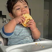 Milán is registered to the contest to win money with this photo: child, toddler, high_chair, bread, eating, crumbs, striped_clothing, indoor, face, hands, happy, food, meal, home, person, cute, baby, furniture, light, expression
