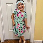 Aitana is registered to the contest to win money with this photo: child, girl, smiling, dress, headband, sandals, door, wooden_floor, indoor, playful_pose, happy, cute, flower_pattern, young, person, footwear, casual_clothing, standing, portrait, home