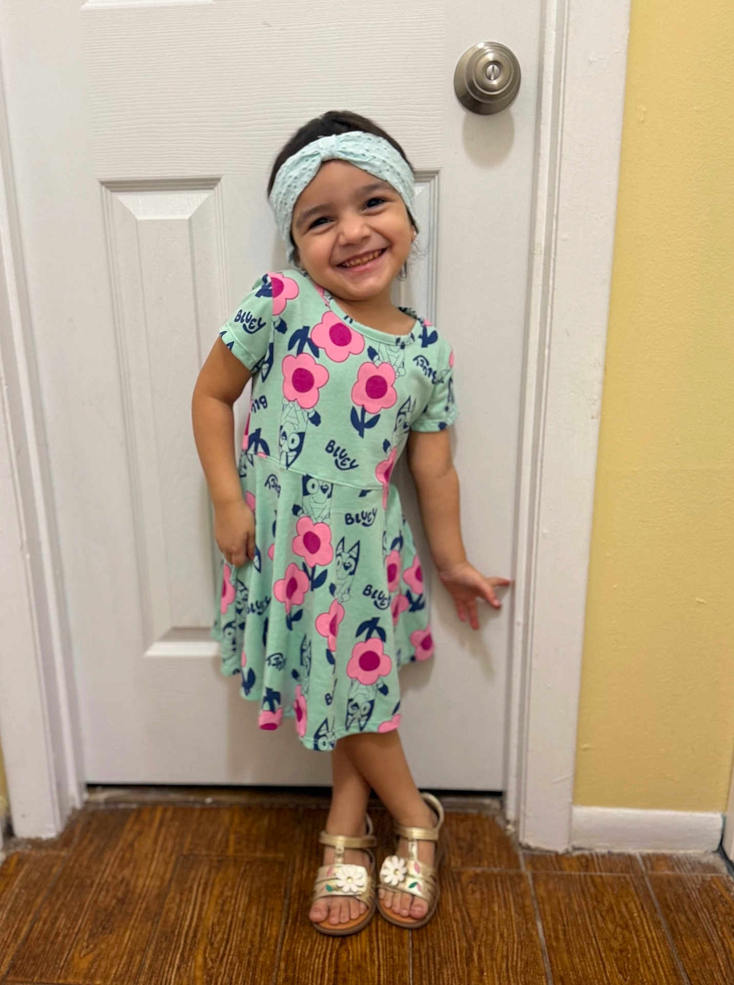 Aitana is registered to the contest to win money with this photo: child, girl, smiling, dress, headband, sandals, door, wooden_floor, indoor, playful_pose, happy, cute, flower_pattern, young, person, footwear, casual_clothing, standing, portrait, home