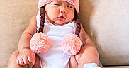 Elara Lucynn joined the competition — help win amazing prizes! baby, child, pink_hat, pom_poms, white_onesie, couch, beige_couch, indoors, cute, sitting, portrait, infant, funny_expression, knitted_hat, plush, cozy, soft_texture, footwear, adorable, young_child