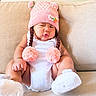baby, child, pink_hat, pom_poms, white_onesie, couch, beige_couch, indoors, cute, sitting, portrait, infant, funny_expression, knitted_hat, plush, cozy, soft_texture, footwear, adorable, young_child