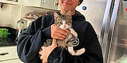 Warren is registered to the contest to win money with this photo: black_hoodie, cabinets, casual_clothing, cat, countertop, domestic, food_items, happy, holding, indoors, kitchen, kitten, mug, pet, portrait, refrigerator, shelf, smiling, woman, young_adult