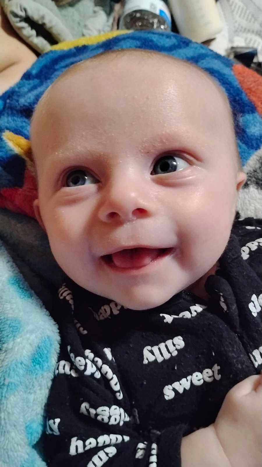 Hudson joined the competition — help win amazing prizes! baby, smiling, close_up, face, blue_eyes, blanket, clothing, happy, infant, child, portrait, cute, soft_texture, indoor, person, black_outfit, text_on_clothing, cheerful, bright, young