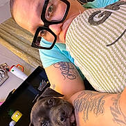 Boolet is registered to the contest to win money with this photo: person, dog, puppy, tattoo, glasses, face, arm, couch, indoor, shelf, curtain, decor, plant, closeup, portrait, casual, snuggle, black_dog, home, cute
