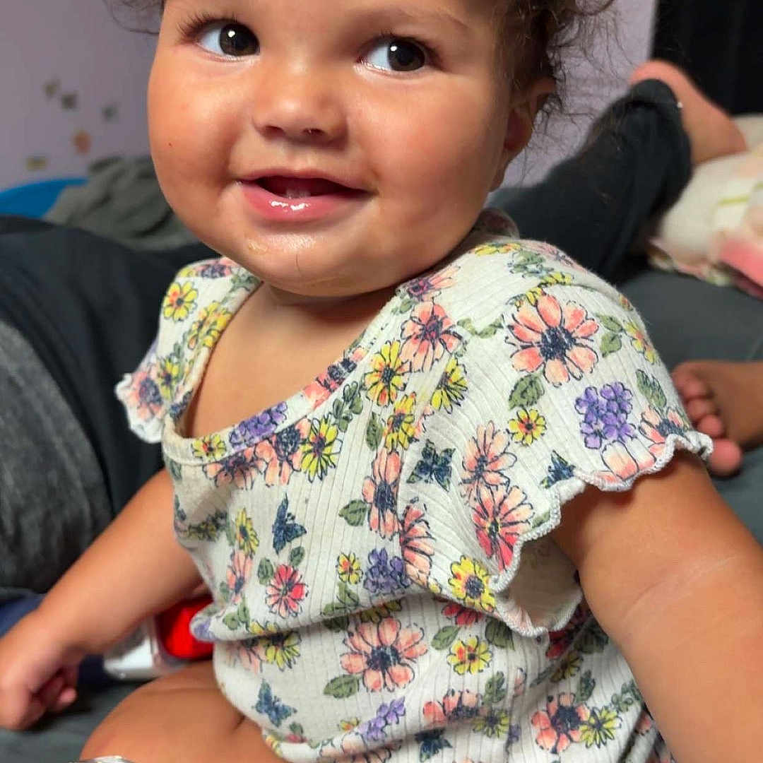 Zyla is registered to the contest to win money with this photo: bed, casual, child, clothing, curly_hair, cute, expression, face, floral_shirt, happy, indoors, person, playful, portrait, sitting, skin, smile, toddler, water_bottle, young_child