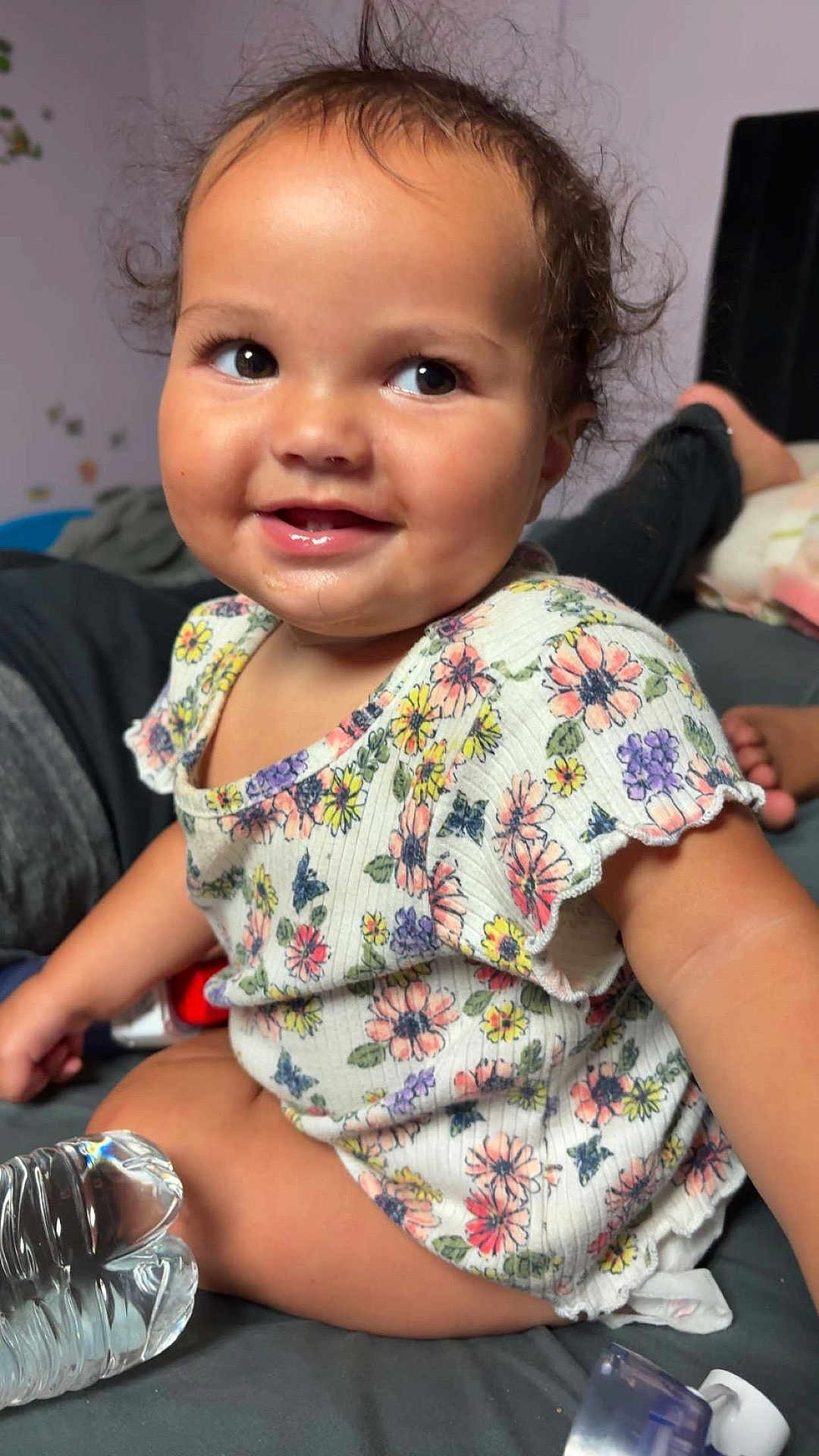 Zyla is registered to the contest to win money with this photo: toddler, child, smile, curly_hair, floral_shirt, sitting, indoors, bed, water_bottle, happy, cute, portrait, face, skin, person, clothing, young_child, playful, expression, casual