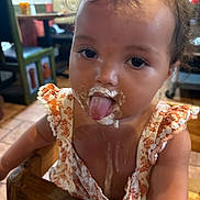 Zyla is registered to the contest to win money with this photo: toddler, child, messy, food, tongue_out, high_chair, indoor, wooden_furniture, dress, curly_hair, face, playful, brown_eyes, table, chair, floor, person, cute, baby, dining_area