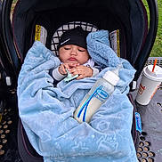 Karsen is registered to the contest to win money with this photo: baby, blanket, car_seat, pacifier, bottle, beanie, outdoor, table, drink_cup, greenery, child, infant, cozy, clothing, person, face, hand, sitting, soft_texture, relaxing