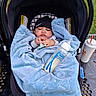 baby, blanket, car_seat, pacifier, bottle, beanie, outdoor, table, drink_cup, greenery, child, infant, cozy, clothing, person, face, hand, sitting, soft_texture, relaxing