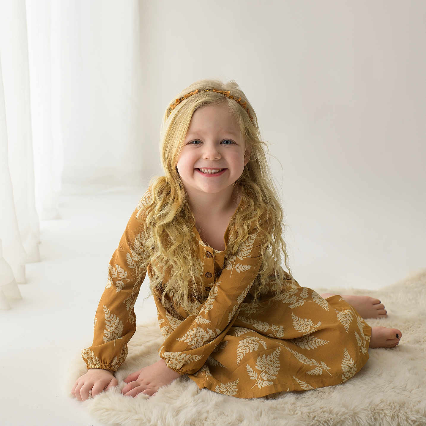 Blakely joined the competition — help win amazing prizes! child, girl, smiling, blonde_hair, curly_hair, dress, mustard_yellow, leaf_pattern, sitting, rug, fluffy_rug, indoor, natural_light, curtains, minimalist, portrait, happy, cute, young, casual