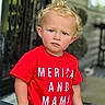 background_blur, blonde_hair, blue_eyes, child, curly_hair, daylight, face, hand_on_chair, outdoor, patterned_pants, person, porch, red_shirt, serious_expression, short_sleeves, skin, standing, text_on_clothing, toddler, young_child