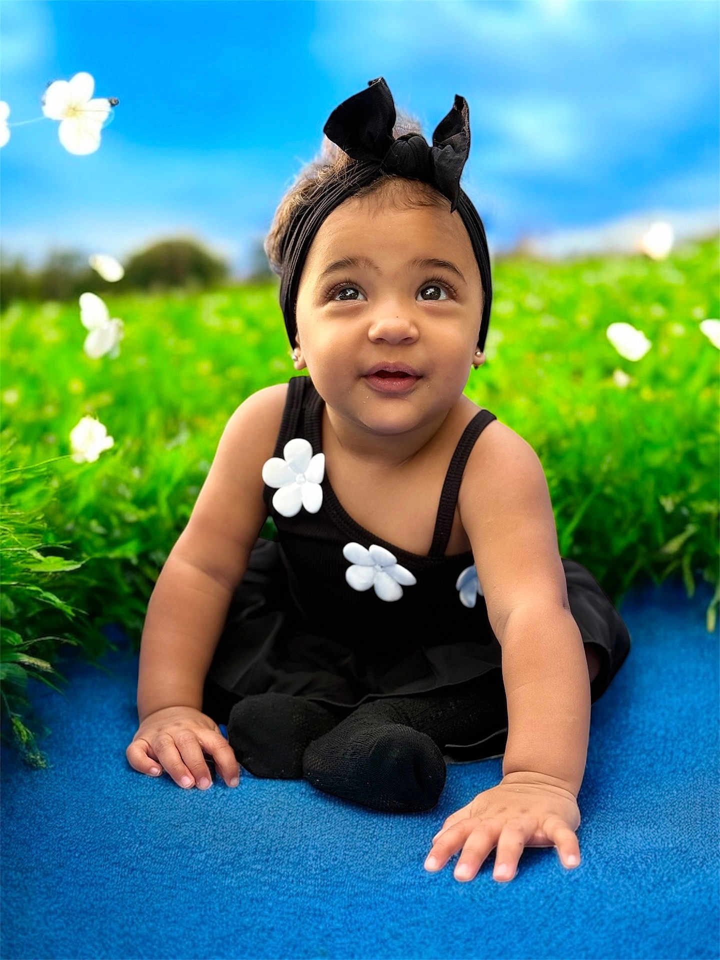 Mia is registered to the contest to win money with this photo: toddler, child, black_dress, headband, flower, grass, blue_surface, outdoor, cute, smiling, baby, nature, greenery, portrait, sitting, daylight, happy, innocent, playful, headwear