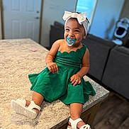Mia joined the competition — help win amazing prizes! toddler, child, green_dress, white_sandals, headband, pacifier, smiling, indoor, marble_countertop, living_room, furniture, happy, cute, baby, person, footwear, accessories, home, portrait, sitting