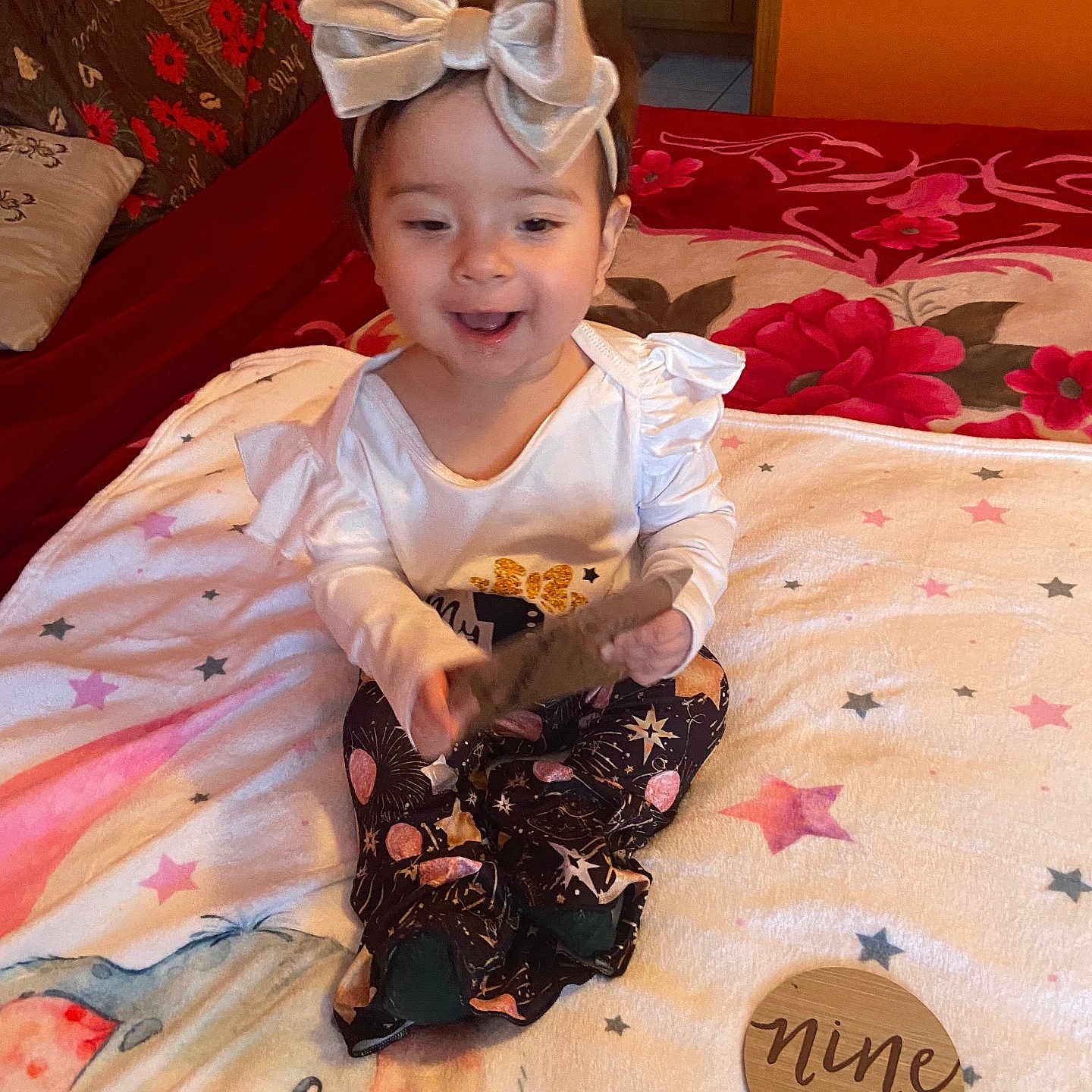 Sophia is registered to the contest to win money with this photo: baby, child, infant, bow_headband, white_top, patterned_pants, blanket, bedspread, elephant_illustration, stars, wooden_plaque, nine_months, indoor, smiling, sitting, cute, happy, portrait, milestone, person