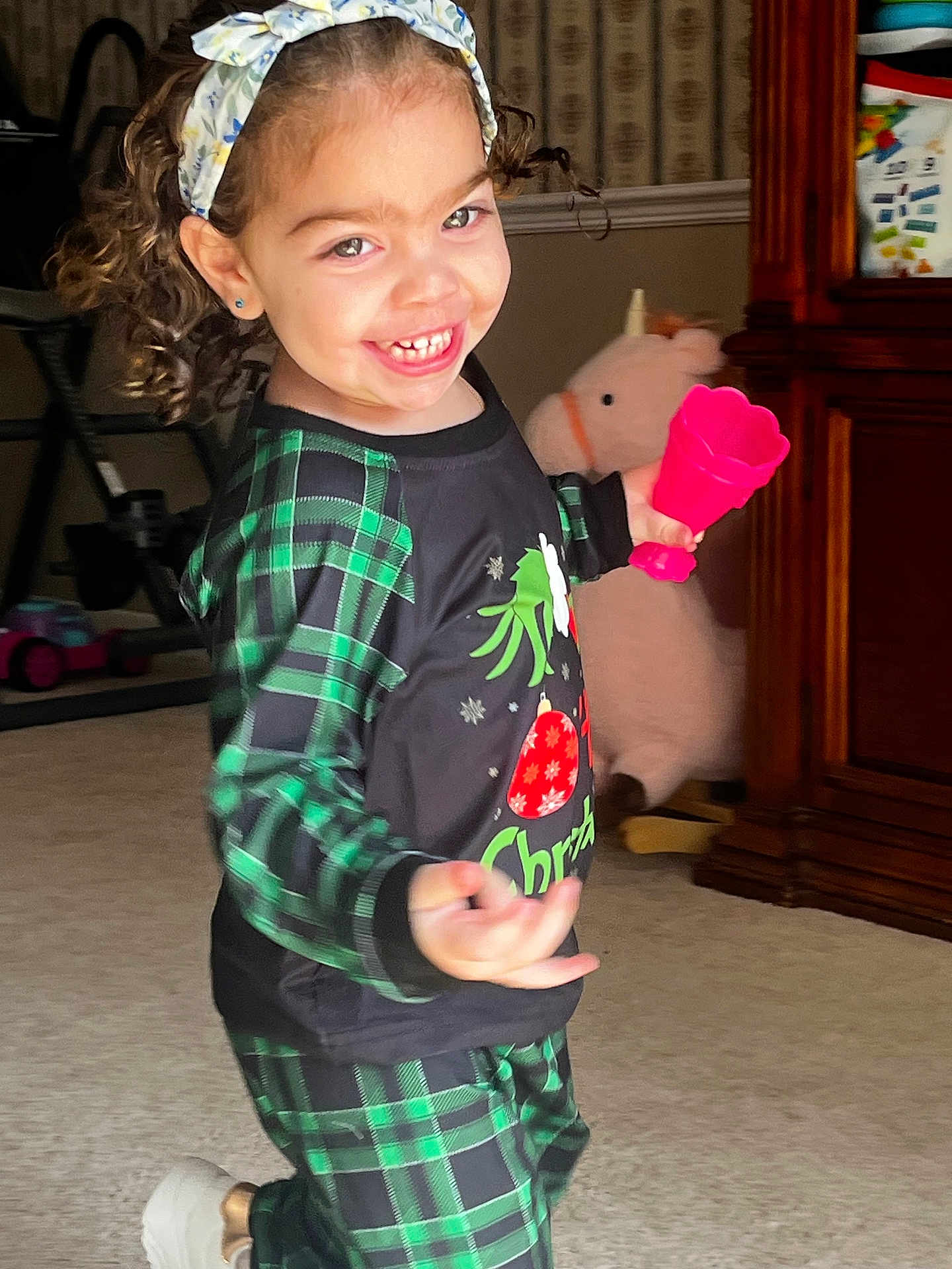 Mila is registered to the contest to win money with this photo: child, toddler, smiling, indoor, plaid_clothing, green, black, toy, pink, headband, curly_hair, carpet, furniture, wooden_cabinet, plush_toy, happy, person, holiday, christmas, play