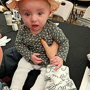 Roselynn is registered to the contest to win money with this photo: baby, child, hat, floral_shirt, white_pants, hand, table, chairs, indoor, person, face, curious, seated, napkin, cutlery, salt_shaker, pepper_shaker, number_sign, dining, casual