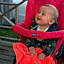 baby, child, stroller, red, orange_blanket, smiling, happy, outdoor, wooden_deck, car, clothing, hands, face, person, cute, infant, seated, hoodie, expression, daylight