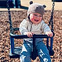 child, baby, toddler, swing, playground, knit_hat, bear_hat, smiling, happy, outdoor, sunlight, wood_chips, swing_chains, play_equipment, hands, jacket, pants, shoes, face, portrait