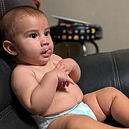 Juan is registered to the contest to win money with this photo: baby, infant, diaper, sitting, black_leather_couch, tongue_out, chubby, cute, indoor, child, face, hands, expression, blurred_background, furniture, pac_man_pattern, stool, skin, portrait, young_child