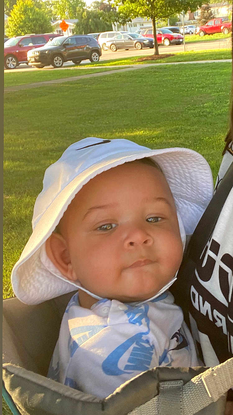 Zamari is registered to the contest to win money with this photo: baby, child, hat, bucket_hat, face, person, outdoor, greenery, grass, park, daylight, car, vehicle, sunlight, smile, carrier, clothing, shirt, closeup, expression