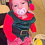baby, infant, child, pacifier, elf_costume, red_clothing, green_clothing, striped_pants, holiday_outfit, indoor, person, face, headwear, sitting, hand, fabric, toy, furniture, bag, floor