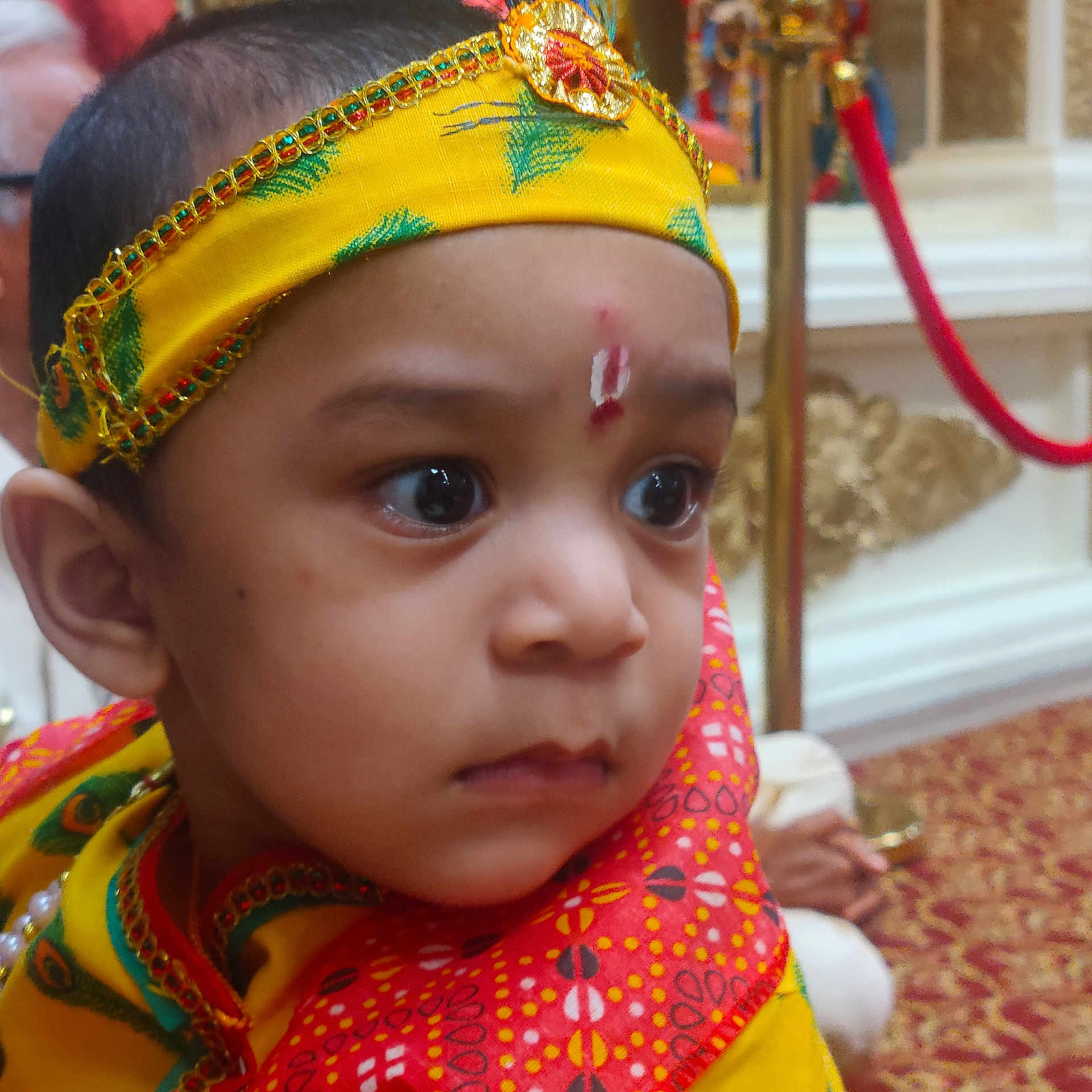 Juhit joined the competition — help win amazing prizes! baby, child, infant, portrait, close_up, face, eyes, wide_eyed, headpiece, traditional_clothing, colorful, yellow, red, patterned_fabric, ceremonial, indoor, decorations, cultural_attire, looking_at_camera, festive