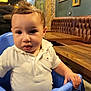 Imran a rejoint le concours — aidez-le/la à gagner de superbes lots ! toddler, child, indoor, high_chair, white_shirt, striped_shorts, curly_hair, restaurant, brick_wall, wooden_table, empty_frames, blue_chair, person, seated, cute, baby, face, hand, casual_clothing, curious