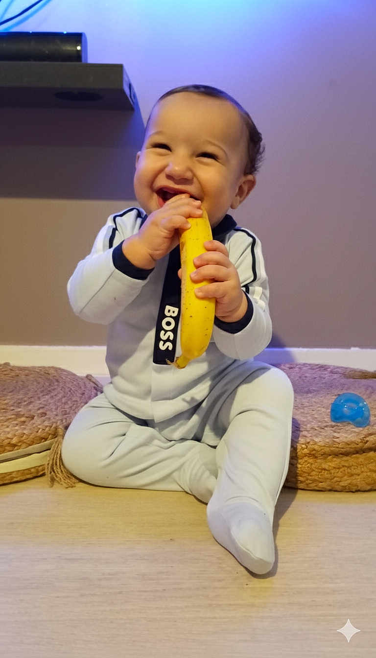 Imran a rejoint le concours — aidez-le/la à gagner de superbes lots ! baby, child, banana, smiling, sitting, onesie, floor, cushion, indoor, happy, food, playful, infant, person, cute, toddler, toy, home, light, portrait