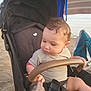 toddler, child, stroller, beach, umbrella, towel, curly_hair, sand, outdoor, sunshade, baby_clothing, seatbelt, hand, leg, sunlight, fence, casual, exploring, summer, relaxation