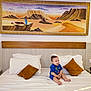 Imran a rejoint le concours — aidez-le/la à gagner de superbes lots ! baby, child, bed, pillow, blue_clothing, white_bed_sheet, wooden_headboard, painting, desert_landscape, mountains, sunset, indoor, smiling, sitting, room, home_interior, wall_art, cozy, happy, infant
