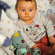 Enéa a rejoint le concours — aidez-le/la à gagner de superbes lots ! baby, child, plush_toy, stuffed_animal, blanket, toy, indoor, curious, face, expression, seated, clothing, striped_shirt, pattern, cushion, nursery, colorful, soft, cute, playtime