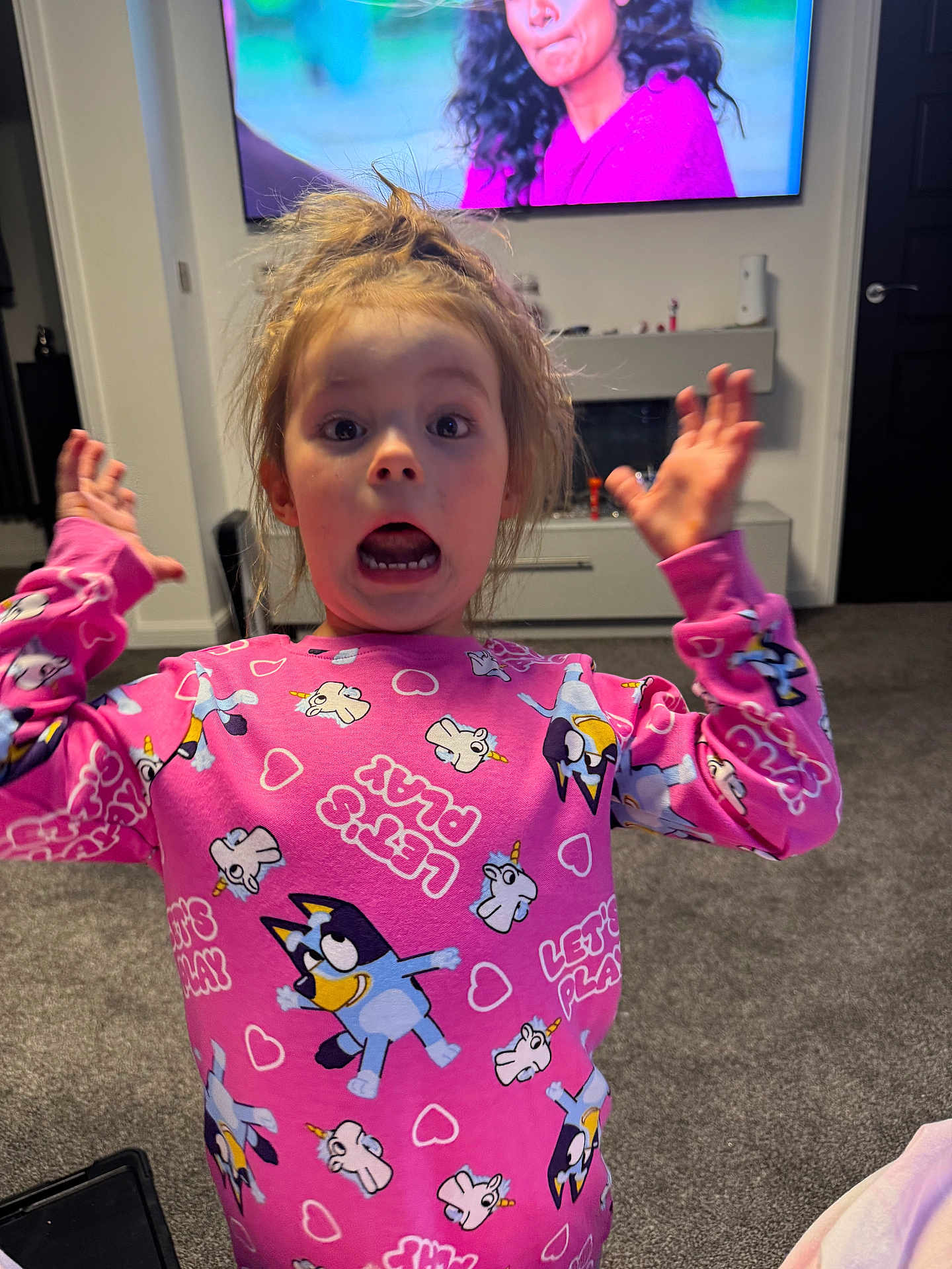 Millie joined the competition — help win amazing prizes! child, surprised, pink_shirt, unicorn, indoor, living_room, television, carpet, furniture, expression, person, young, casual_clothing, home, cute, funny_face, hands_up, hair_up, carpeted_floor, wall