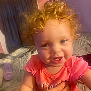 Nakiya is registered to the contest to win money with this photo: baby, child, infant, smiling, curly_hair, portrait, indoors, bedroom, pink_clothing, coral_shirt, happy, face, eyes, teeth, sitting, adult_hand, family, blanket, bed, cozy