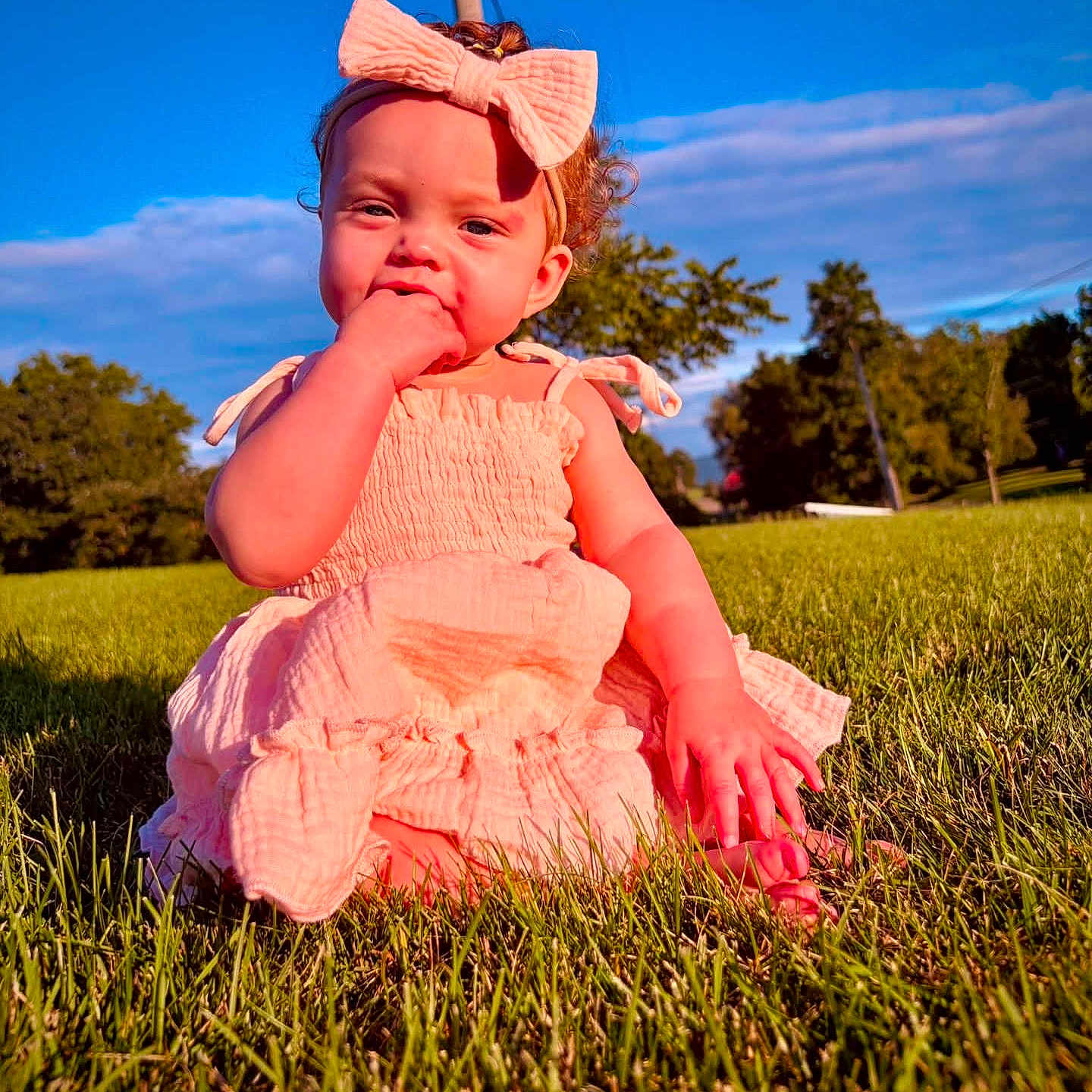 Nakiya is registered to the contest to win money with this photo: baby, child, infant, outdoor, grass, park, portrait, sitting, bow, headband, dress, pink_clothing, smiling, hand_to_mouth, curly_hair, face, sunlight, blue_sky, trees, nature