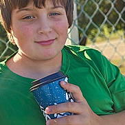 Elisha joined the competition — help win amazing prizes! child, boy, green_shirt, cup, outdoor, sunlight, fence, hand, portrait, smile, casual, daylight, young, holding, blue_cup, background, nature, person, relaxation, park