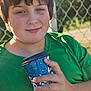 child, boy, green_shirt, cup, outdoor, sunlight, fence, hand, portrait, smile, casual, daylight, young, holding, blue_cup, background, nature, person, relaxation, park