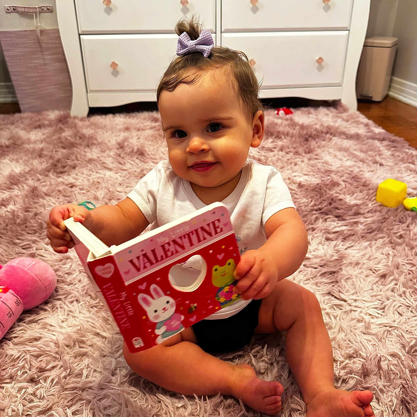 Alaiah joined the competition — help win amazing prizes! baby, book, child, clothing, cute, decor, feet, floor, hand, indoor, infant, pink_rug, playful, purple_bow, room, sitting, smiling, toy, valentine, white_dresser