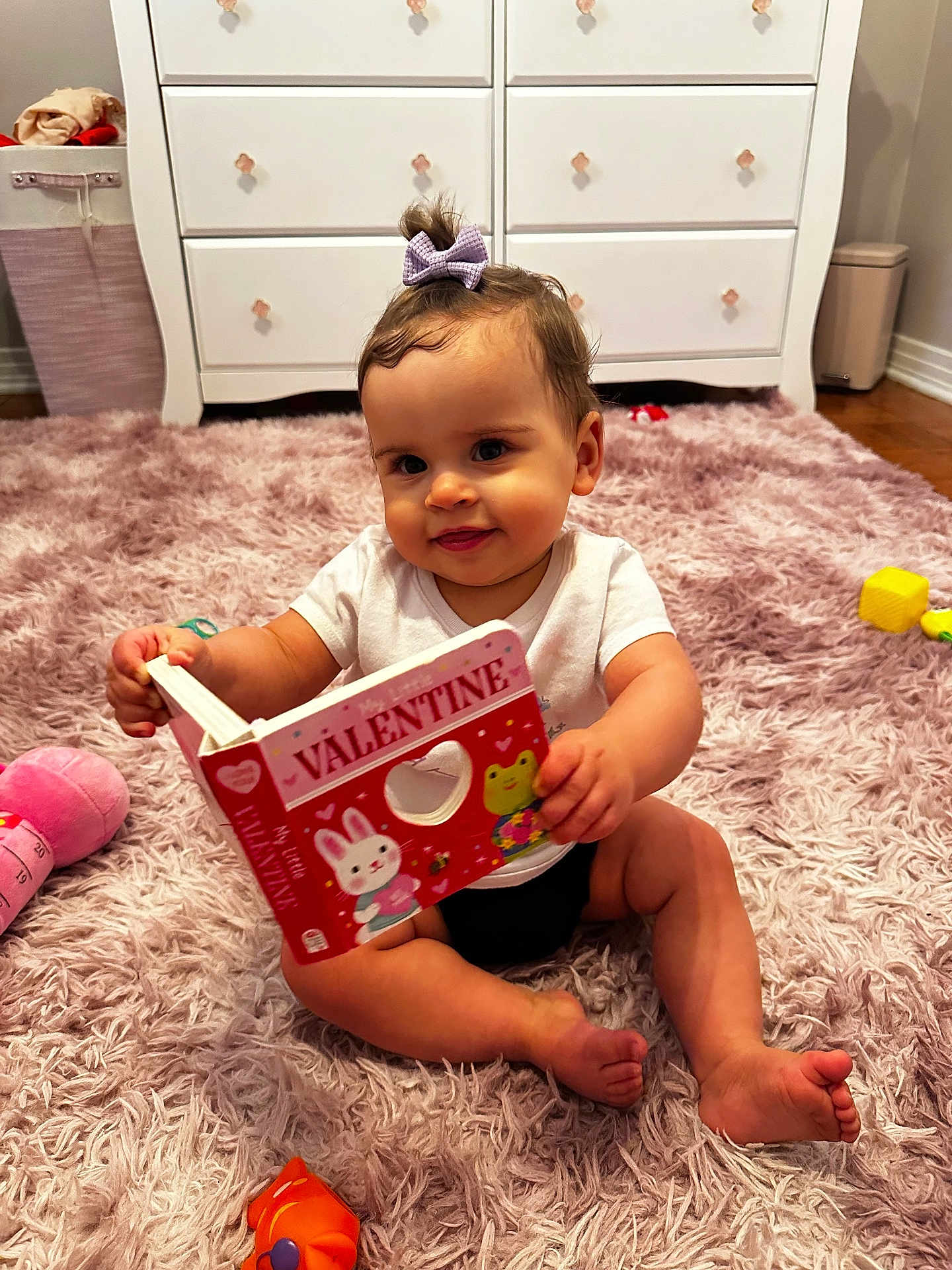 Alaiah joined the competition — help win amazing prizes! baby, child, book, valentine, toy, pink_rug, purple_bow, white_dresser, floor, smiling, sitting, indoor, cute, infant, playful, decor, feet, hand, clothing, room
