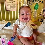 Raelynn is registered to the contest to win money with this photo: baby, smiling, hospital_bed, medical_tubes, bow, infant, child, hospital_room, medical_equipment, person, cute, healthcare, happy, sitting, bandage, white_clothing, orange_shorts, face, skin, indoors
