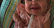 Raelynn is registered to the contest to win money with this photo: baby, smiling, infant, pink_dress, hair_accessories, medical_tube, cushion, striped_pattern, happy, child, indoors, cute, healthcare, comfort, person, portrait, sitting, young_child, expression, closeup