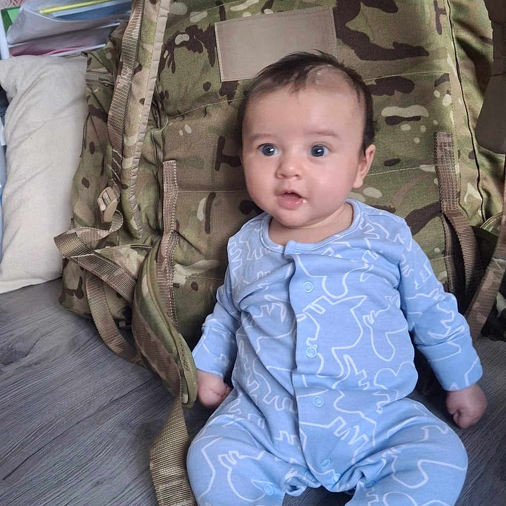 Cooper joined the competition — help win amazing prizes! baby, backpack, blue_onesie, camouflage, child, clothing, curious_expression, cute, face, floor, household_items, indoor, infant, person, pillow, shelf, sitting, wide_eyes, wooden_floor, young