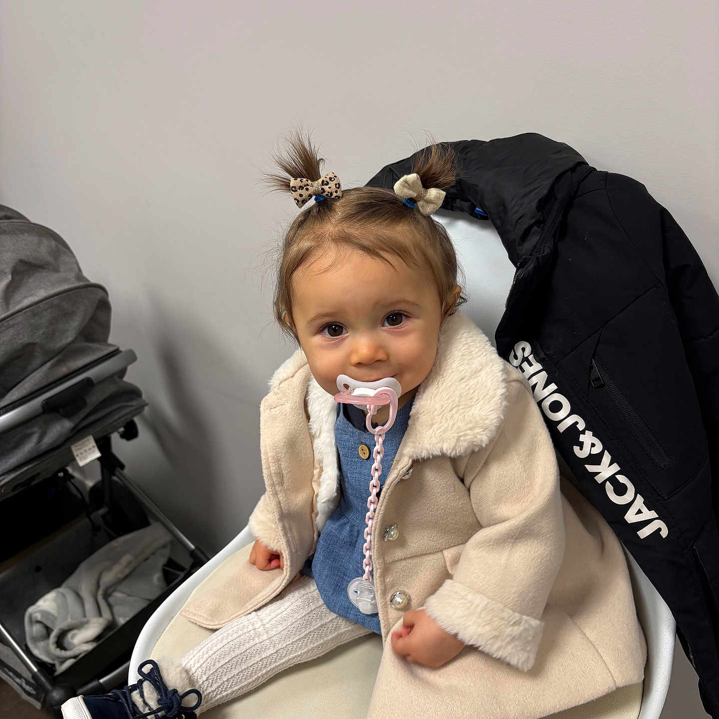 Tahyssia participe au concours pour gagner de l'argent avec cette photo : accessory, baby, boots, bow, chair, child, clothing, coat, cute, indoor, jacket, knitted_leggings, pacifier, person, pigtails, sitting, smiling, stroller, toddler, white_wall