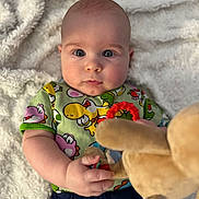 Emanuel is registered to the contest to win money with this photo: baby, bed, blanket, blue_eyes, cheeks, child, close_up, colorful_clothing, cozy, cute, face, gaze, hand, infant, lying_down, onesie, plush_toy, portrait, soft_texture, toy_ring