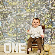 Maddox joined the competition — help win amazing prizes! toddler, child, birthday, celebration, chair, teddy_bear, smiling, one_year_old, white_shirt, brown_pants, floor, sign, happy, indoors, portrait, festive, decorations, chalkboard, colorful, wooden_chair