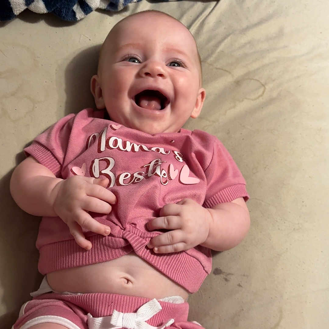 Kennedy is registered to the contest to win money with this photo: baby, infant, smiling, pink_clothing, shorts, tshirt, blanket, striped_blanket, lying_down, happy, cute, child, person, indoor, skin, head, arms, legs, belly, casual