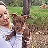 woman, person, puppy, dog, pit_bull, pet, animal, portrait, outdoor, park, grass, tree, playground, play_equipment, holding, sitting, brown_dog, brown_hair, casual_clothing, nature