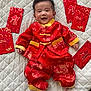 baby, smiling, red_clothing, traditional_outfit, quilted_blanket, red_envelopes, celebration, festive, infant, portrait, happy, cultural, indoors, fabric, patterned_clothing, person, cute, lying_down, asian_baby, decorative